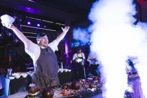 A chef raises his arms as smoke rises in front of a dessert smash he is preparing on a table in front of him