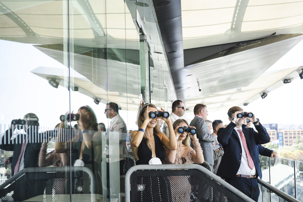 A group of people with binoculars watch a horse race from the balcony of a grandstand