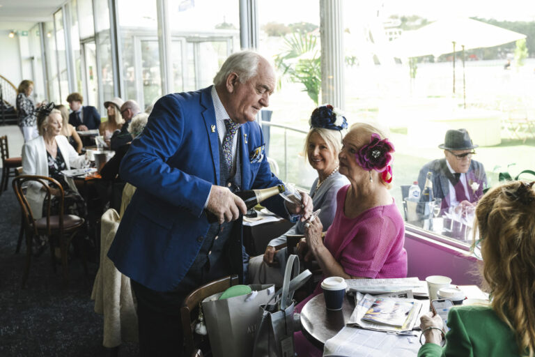 An elderly man pours champagne for his wife in a restaurant at a racecourse