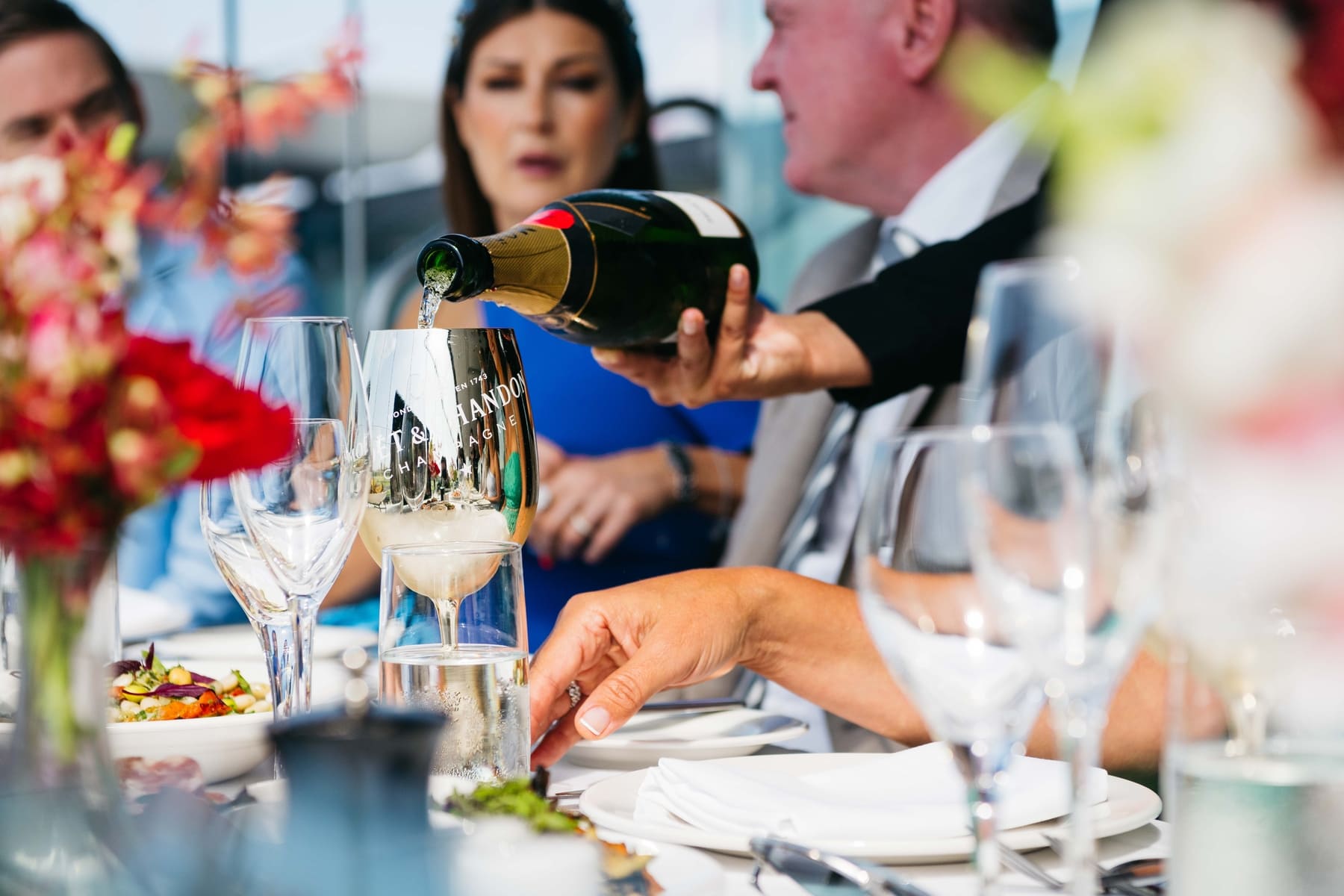 A group of racegoers sit at a dining table as champagne is poured.